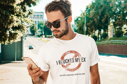 Man wearing "I Blocked My Ex" graphic tee, looking at his phone while standing on a sunny city street.