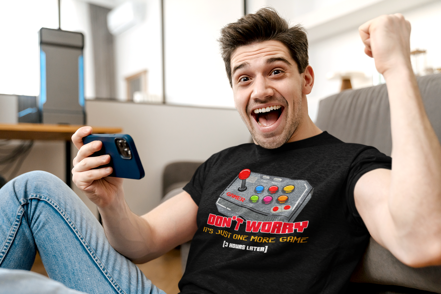 Man cheering on couch wearing black "Just One More Game" gamer T-shirt while holding a phone
